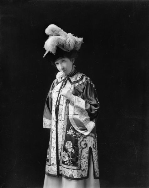 The photographer's wife, dressed in embroidered oriental jacket and plumed hat, stands holding a rose.