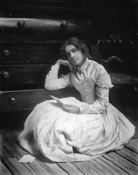 A young woman sits on the floor, one arm resting on an open dresser drawer, gazing into the distance and holding a letter. She is wearing an old-fashioned dress.