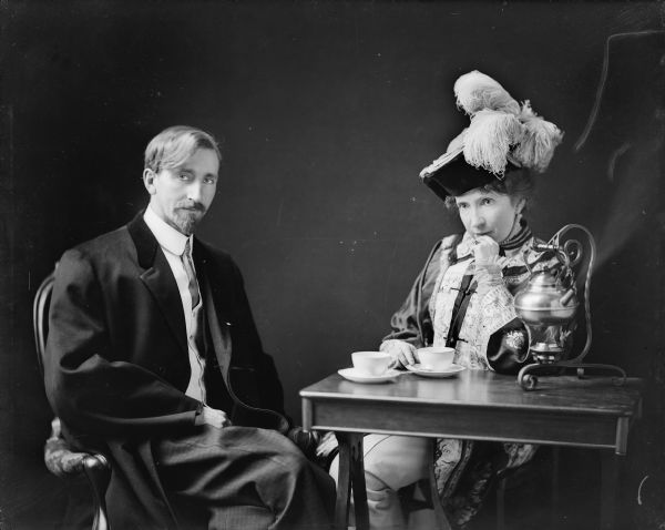 The photographer, Ephraim Burt Trimpey, and his wife, Alice Kent Trimpey, seated at a table with a tea kettle on a stand, cups and saucers. Both wear vintage costumes; Mrs. Trimpey's includes a plumed hat and oriental jacket.