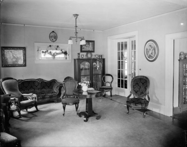 An interior view of a parlor or sitting room, possibly the photographer's, with furnishings of several periods and styles. There is a Victorian settee and chairs, Gothic Revival bookcase, and Arts and Crafts Style light fixture.