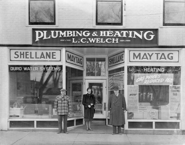 Two men and a woman pose in front of the L.C. Welch Plumbing and Heating store, 137 Third Avenue. There are signs advertising Maytag and Shellane, "compressed natural gas." A Kohler model bathroom is in the left display window; kitchen stoves are in the right.