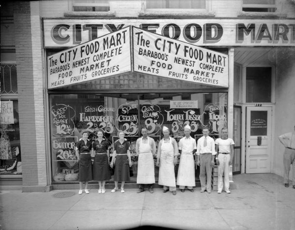 Five men and three women, employees of City Food Mart, pose in front of the store at 124 Third Street. Signs identify the store as "Baraboo's Newest Complete Food Market." There are watermelons in the front window.