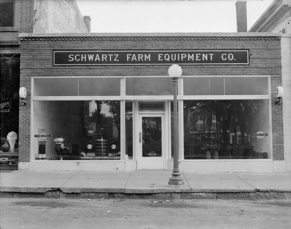 Exterior from street of the Schwartz Farm Equipment building, 135 Third Avenue. Show windows in the one-story brick building feature an International truck on the left and a Farmall tractor on the right.  There is a cream separator in the left display window.
