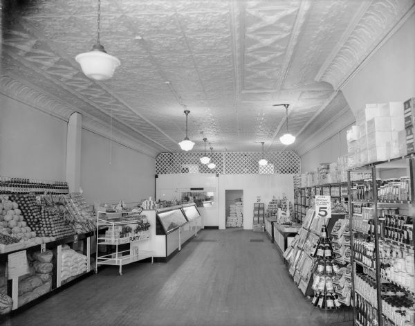 The interior of a small food market with produce, beverages, canned goods and coolers. This appears to be the City Food Mart. There is a decorative pressed tin (steel) ceiling and wood floor.