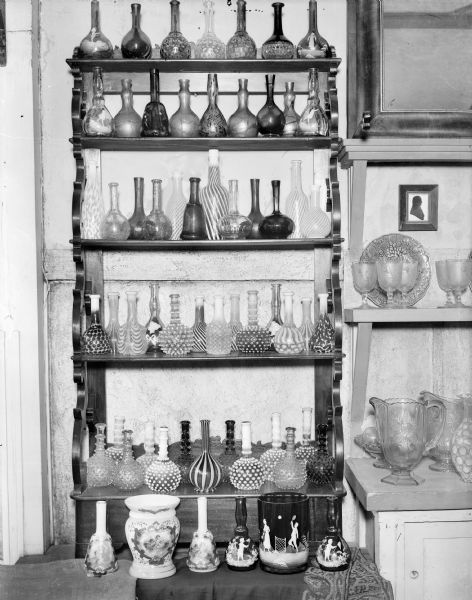 A display of glass barber's bottles in the home of the photographer.  There are examples of blown glass in a variety of painted, iridescent, and hobnail designs. These bottles, with dispensing stoppers, were once common in barber shops. In 1906, however, the Pure Food and Drug Act made it illegal for barbers to refill bottles of lotions and tonics, making these bottles obsolete.  On the lowest shelf are two sets of barber's bottles with matching waste "vases."