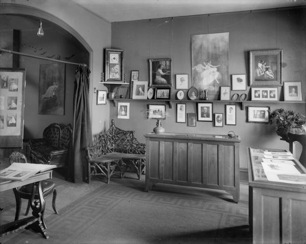 A room in the home of Ephraim Burt and Alice Kent Trimpey. The walls are decorated with photographs, paintings, and prints. There are two glass-topped wooden display cases and a corner settee in the rustic style. A large bare light bulb hangs in the arched entrance. The carpet, probably sisal, has a Greek key border.
