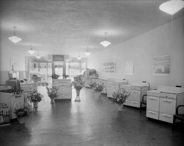 An interior view of Pointon Appliance, 132 Fourth Avenue, showing a variety of Maytag washers, kitchen ranges, and water heaters. There are signs for Shellane bottled gas and gas refrigerators. There are several floral arrangements throughout the store, possibly on the occasion of the store's grand opening January 19, 1946. The Sauk County Courthouse and snow-covered square are visible through the front windows.