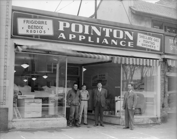 Four men pose in front of the Pointon Appliance store, 132 Fourth Avenue, located between the Al. Ringling Theatre and Trimpey Studio.  Signs advertise Frigidaire, Bendix Radios, Maytag washers and Shellane Gas. Several appliances are displayed in the front windows. The Sauk County Courthouse is reflected in the left front window. The store opened January 19, 1946.