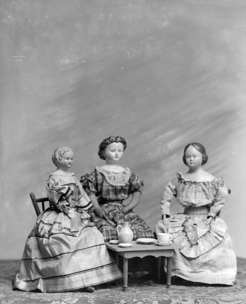Three dolls from the collection of the photographer's wife, Alice Kent Trimpey, are posed seated at a small table with a tea set. The dolls date from the mid-nineteenth century; the doll on the left was known as "Miss Luciny."