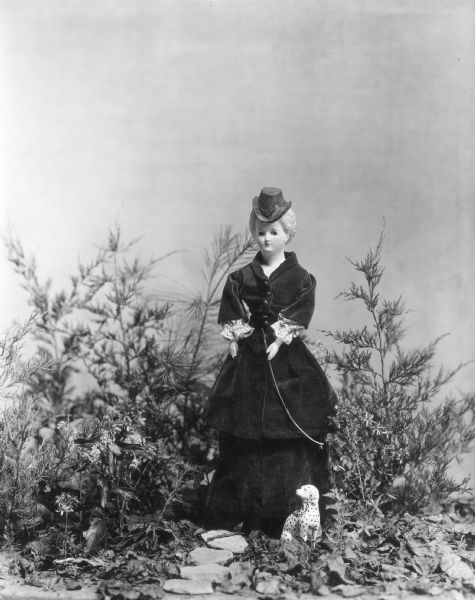 A doll from the collection of Alice Kent Trimpey and dating to the mid-nineteenth century is posed in a woodland setting with a small china dog. The doll's head and shoulders are wax over papier-mache.  She wears a velvet riding habit and hat and carries a crop.