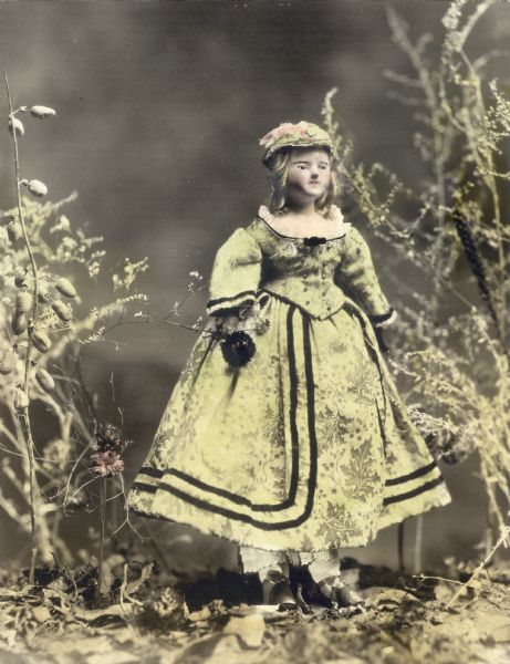 Hand-colored photograph of a doll, Martha, from the collection of Alice Kent Trimpey, posed in a woodland setting. She wears a green brocade dress and hat. Her head and shoulders are wax over papier mache.