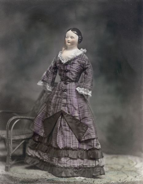A doll with china head from the collection of Alice Kent Trimpey is posed by a miniature chair.  She wears a silk dress and dates from the mid-nineteenth century.
