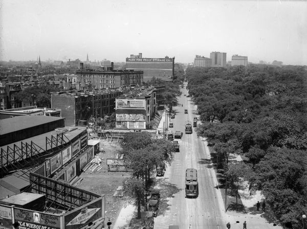 Elevated view north along Clark Street with pedestrians, street cars, trucks and automobiles. Lincoln Park is on the right; the Belden Stratford Hotel is on the right in a group of three large buildings in the background. Advertising signs surround a construction site in the foreground where men are working.
