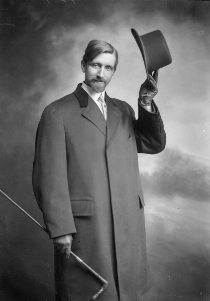 The photographer doffs his hat and smiles at the camera. He is wearing a long coat and gloves, and carries a cane.