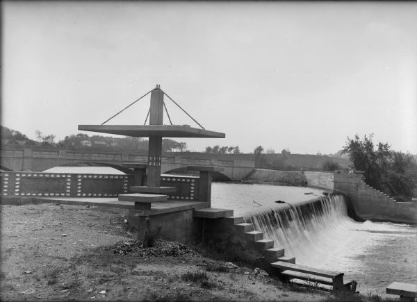 The Island Woolen Mill dam and bridge over the Baraboo River. In the foreground is a concrete "pergola" which shades a small bench. Molded into a concrete wall is the inscription "Erected by the Island Woolen Company 1913." Clamshells have been used as decoration on the wall and pergola.