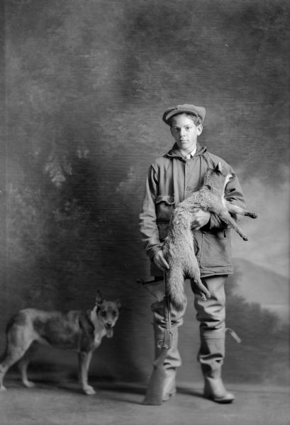 A studio portrait of a young man in front of a painted backdrop wearing rubber boots holding his gun and a dead fox with his dog standing nearby.