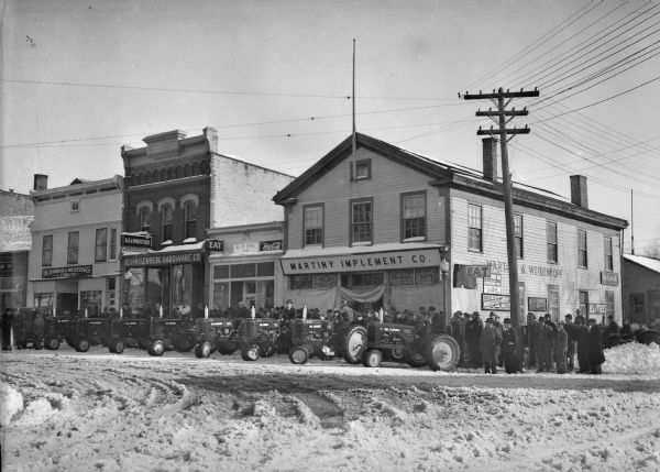 Eight new Massie-Harris tractors are parked in the snow in front of the Martiny Implement Co., 147 Third Avenue. A crowd has gathered and men sit on the tractor seats. Each tractor has a name painted on it, including Mr. Fleming, Mr. Morley, Mr. Marshall, Mr. Thomas, Mr. Mather, Mr. Beardsley and two others. Other businesses shown include the Blue Bird Inn, Bohn-Isenberg Hardware Co. and L.C. Welch Plumbing & Heating.