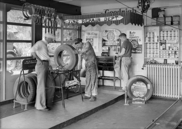 A repair man checks a tire on a stand and a second inspects an inner tube while a customer looks on. Signs advertise Goodrich Tires, Bowes Seal Fast, and there are displays of belts, tires and batteries.