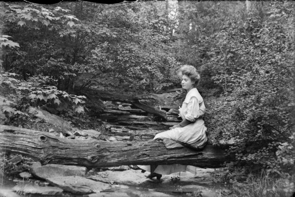 A young woman sits on a log which lies over a stream bed in a wooded area.