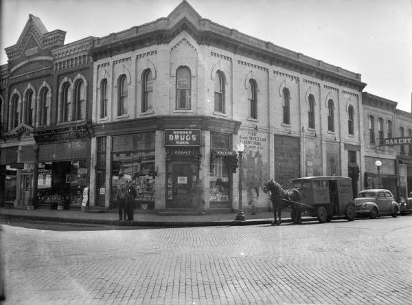 View from cobblestone street of a man standing next to a police officer at the corner of Third and Oak Streets, in front of the Corner Drugstore. A horse-drawn milk wagon with rubber tires is parked on Oak Street. Signs on the drugstore advertise Devil's Lake, Kodak film, and the office of Dr. Tryon, on the second floor. The Atlantic and Pacific (A & P) Tea Company store is next door, on Third Street.