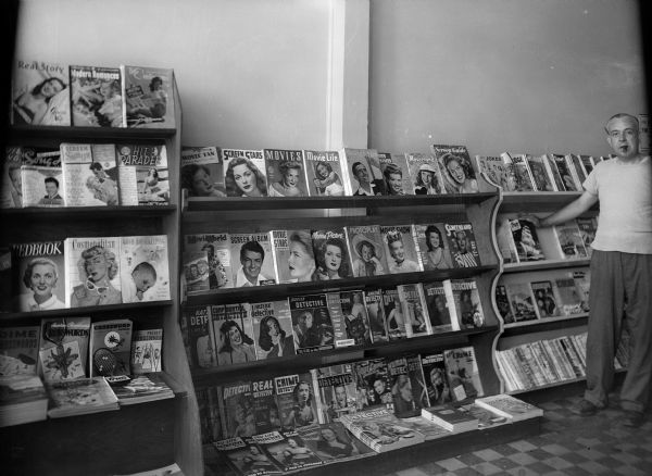 An unidentified man with a cigar stands next to a rack displaying periodicals, including a large number of movie fan and detective magazines.