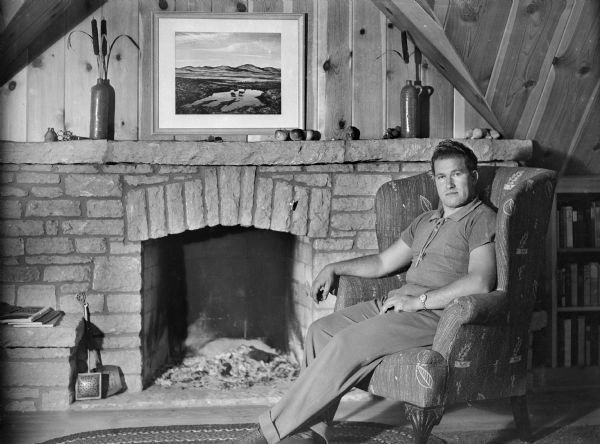 Sauk City author August Derleth sitting in a wing back chair near a fireplace in his home, "The Place of Hawks."