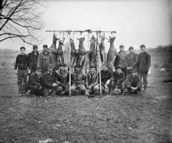 A group of 14 hunters poses in front of seven field dressed deer which hang from a rack made of metal pipe. There is a barn in the far background, left, and two chickens on the grass on the right.