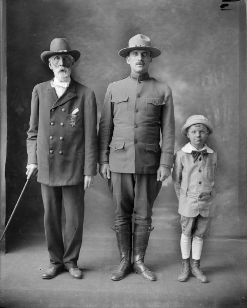 A studio portrait, presumably of Brad Robinson, his son and grandson.  The eldest wears a badge of the Grand Army of the Republic, identifying him as a Civil War veteran. His son, in uniform, has a collar insignia of the United States Reserve Officers; that insignia was used during the first half of the World War I.