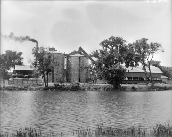 Two large clay block silos stand beside a river. There is a large building, possibly a mill, behind the silos, a barn to the right and a shed on the left. Men pitch hay from wagons into the shed as a horse hitched to one wagon eats from a wagon in front. There is a large smokestack in the background.