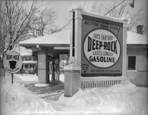 A large sign identifies the Deep Rock service station at 132 Fifth Avenue. Another sign advertises "Kant-Nock" gasoline. There are large piles of snow lining the drives and walk. The stucco-covered building with deep overhangs has two gas pumps.