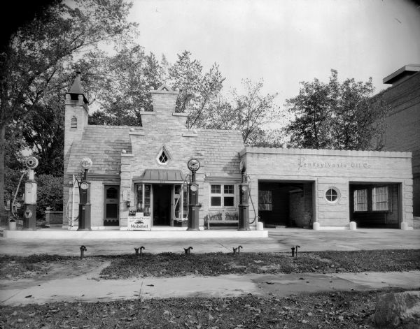 A highly detailed, brick, storybook style Pennsylvania Oil Co. gas station with a slate roof. There is a large chimney on the left and several gas pumps. Men's and women's restrooms are marked, and there is a bench in front. Two service bays are on the right. A display of Mobiloil sits in front.