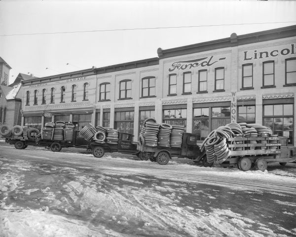 Five trucks loaded with tires are parked in front of the Philbrick Garage, a Ford and Lincoln dealership at 207 Third Avenue. Boxes on the far right truck are marked "Firestone." The garage occupies several older two-story brick buildings. There is snow on the street.