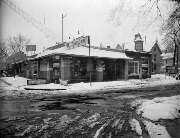 View from street of a cement block building housing an Energee brand gas station and Ben Doty's Tire Service. Next door on the left is Mueller's Garage, a Hudson-Essex dealership, with several automobiles parked in front. To the right, partially obscured, is a two-story brick Victorian house with a turret.  There is snow on the ground.