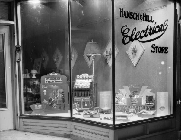 A night view of the window display in the Hansch and Hill Electrical Store, 121 Fourth Avenue. Floor lamps and wall sconces are featured, as are displays of Westinghouse Mazda Lamps (lightbulbs) and electric appliances, including a percolator, iron, toaster, and waffle maker.