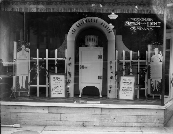 A window display at the Wisconsin Power and Light Company office, 116 Fourth Avenue, features a General Electric refrigerator as "The Gateway to Health" under an arch in a picket fence.