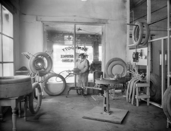 A tire repairman in coveralls and necktie is shown in his shop among tires, inner tubes and equipment. Gas pumps are visible through the front window.