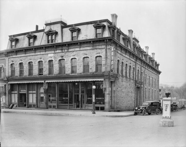 Cars are parked along the side of the Warren Hotel, 102 Fourth Avenue at the corner of Oak Street. The hotel is a large two and one half-story stone structure with mansard roof. A stone above the central second story window bears the year 1877. The dining room occupies a storefront on the first floor on the left. A monument in the intersection marks Wisconsin Highway 12.