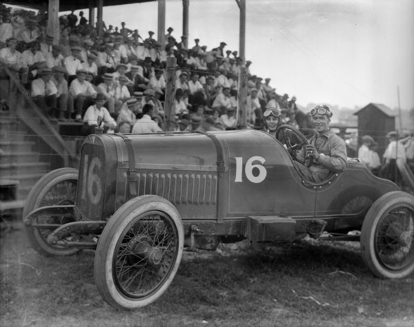 A race car driver and passenger sit in their racer, No. 16, in front of a grandstand. Both wear goggles; the passenger wears a leather helmet. The spectators, nearly all men and boys, wear straw hats or caps.