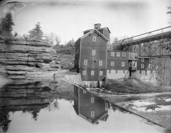A view of the Timme Mill and its reflection in Dell Creek at Mirror Lake. There is a bridge on the right; a wooden building with cupola stands in the background.