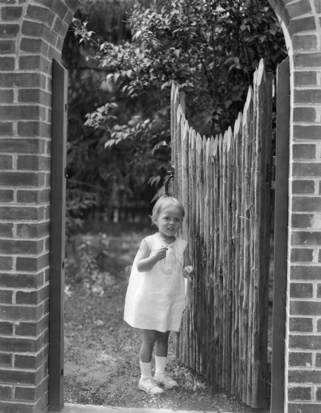 A small child stands by a rustic wooden gate in a brick archway at the home of William Llewellyn and Zona Gale Breese. The child may be Leslyn, the adopted daughter of the Breeses. Zona Gale had assumed custody of Leslyn prior to marrying William Breese.
