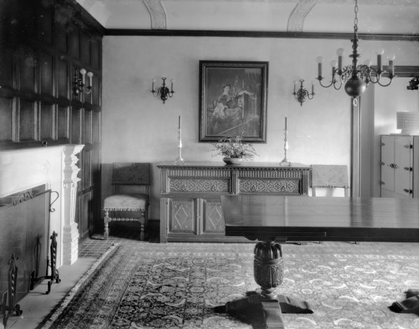 The dining room in the home of William Llewellyn and Zona Gale Breese.  There is a large fireplace, oriental rug, and matching chandelier and sconces. A large refrigerator is visible through the doorway.