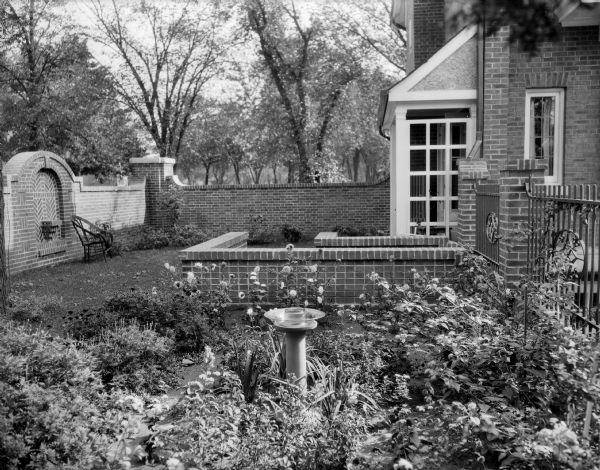 A garden in late summer with dahlias and marigolds at the home of William Llewellyn and Zona Gale Breese. The house, a brick wall and decorative fence enclose the garden. There is a small bird bath and a rustic chair in the garden.