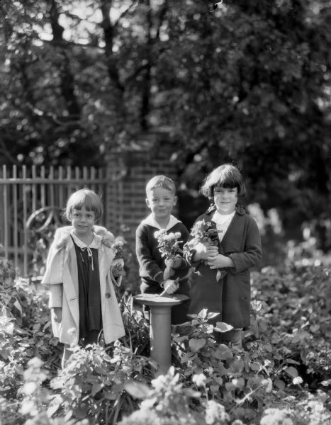 Two girls and a boy pose holding flowers in the garden of William Llewellyn and Zona Gale Breese. The boy grips the gnomon of the sundial. The girl on the left is probably Leslyn, the adopted daughter of the Breeses.