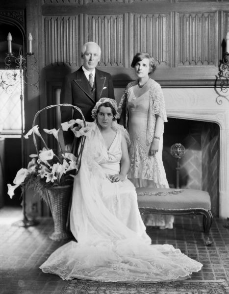 A portrait of the bride, Juliette, seated, with her father, William Llewellyn Breese, and step-mother Zona Gale Breese, standing behind her. There is an arrangement of calla lilies and other lilies in a large basket. The photograph was taken in Zona Gale's study.