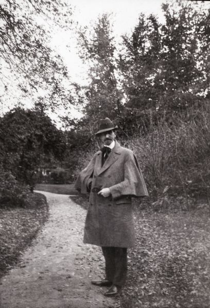 The photographer stands on a park or garden path holding his gloves. He wears a coat with attached cape, hat, and large cravat.