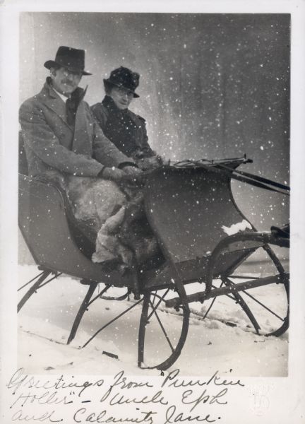 Winter scene with Ephraim Burt and Alice Kent Trimpey seated in a sleigh wearing overcoats and hats, and covered with a fur lap robe. A handwritten message reads "Greetings from 'Punkin Holler' - Uncle Eph and Calamity Jane." There is a small amount of snow on the ground, and the negative was altered to further create a snowy effect. The photographer's initials are embossed in the lower right corner.