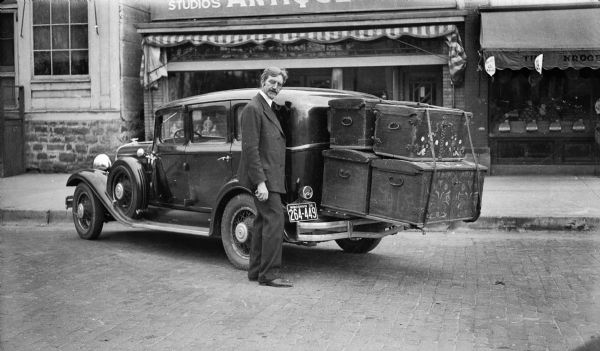 The photographer stands beside an automobile with a 1935 Wisconsin license plate. Four large (possibly Norwegian) trunks with decorative painting are tied to the rear bumper. The car is parked in front of the Trimpey Studios and Antique Shop, which is identifiable by the distinctive Prairie Style door. Dolls in the shop window are visible through the windshield of the car, and belong to the photographer's wife, Alice Kent Trimpey. Produce is visible in the Kroger's Grocery store window to the right.