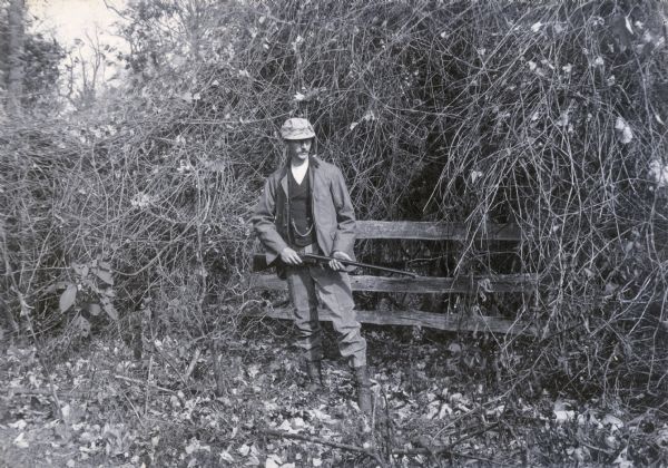 Ephraim Burt Trimpey poses while hunting, holding a shotgun.  He wears a hunting jacket and cap, corduroy pants and high boots.  He also sports a vest with watch chain.