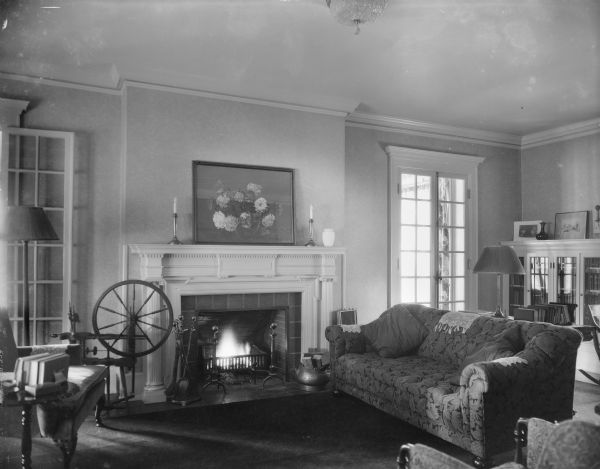 The living room in the home of William Llewellyn and Zona Gale Breese, with a fire in the fireplace. There is a spinning wheel near the hearth and bookcases along the far wall. The sunroom is visible through the French doors.