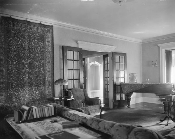 A view of the living room in the home of William Llewellyn and Zona Gale Breese, looking toward the front entry hall. There is a grand piano in the front of the room. A large oriental rug hangs on the wall; a lamp with a stained glass shade sits on a table beside a chair.
